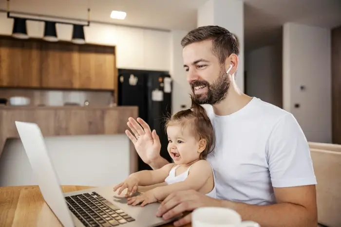 A happy casual businessman is having conference call with coworkers and greeting them while his daughter in his lap is helping him with work.