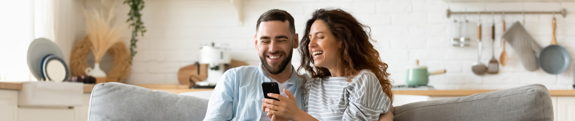 Happy young woman and man hugging, using smartphone together, sitting on cozy couch at home